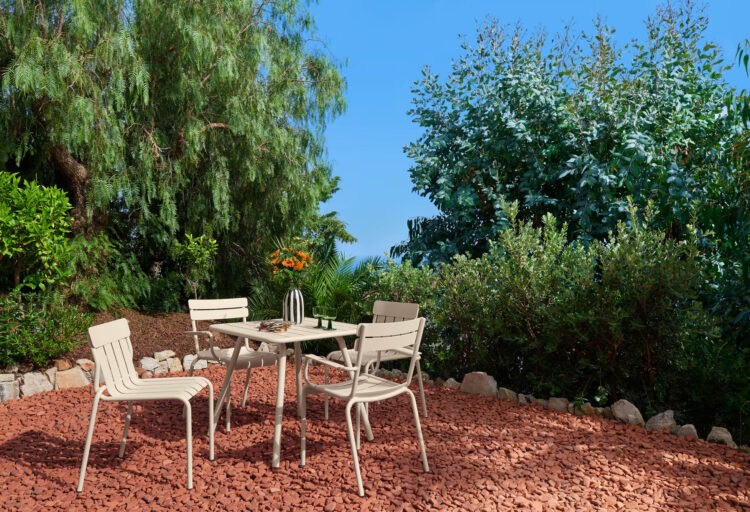 Outdoor dining set in Latte Beige by Fermob sits on red gravel under lush greenery, the warm neutral outdoor furniture finish softening the landscape while framing a relaxed garden setting.