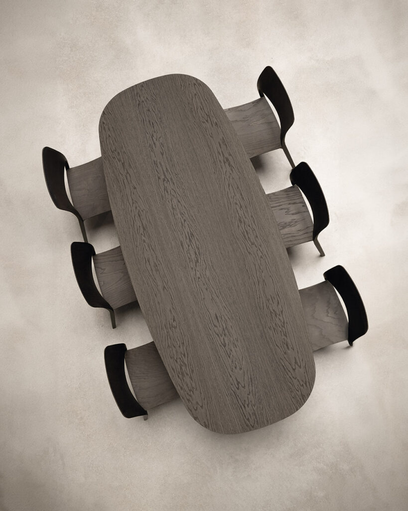 Oval pedestal table in grey oak with petal-shaped plywood base, surrounded by black dining chairs, viewed from above.