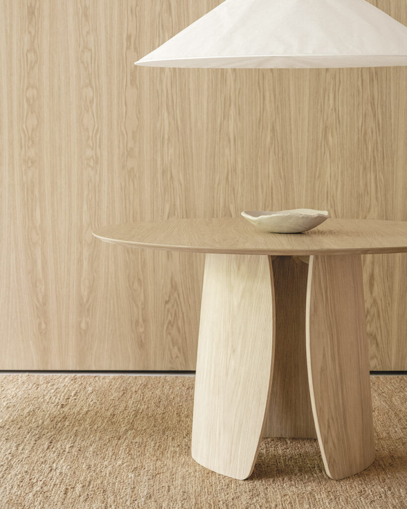 Round oak pedestal table with curved plywood petal base beneath a soft pendant light in a minimalist wood interior