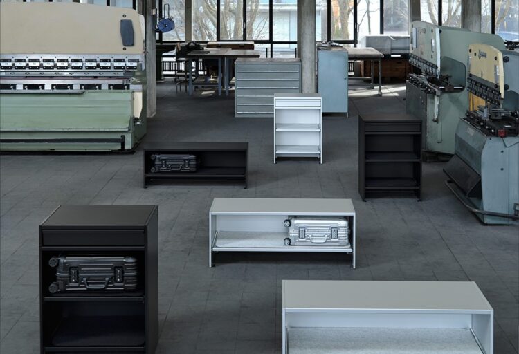 Assorted aluminum storage furniture units in a factory setting, benches and cabinets holding suitcases like curated objects.