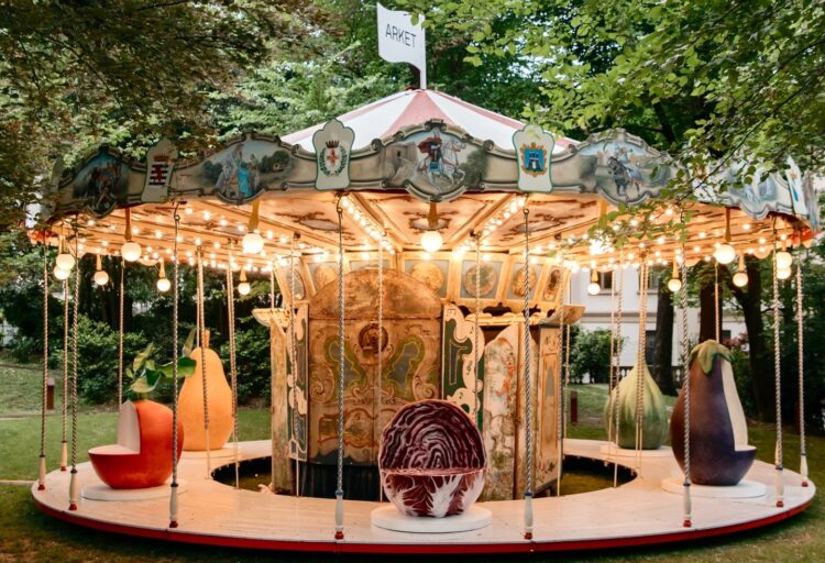 Restored antique carousel surrounded by trees, topped with an Arket flag and filled with oversized fruit sculptures as seating