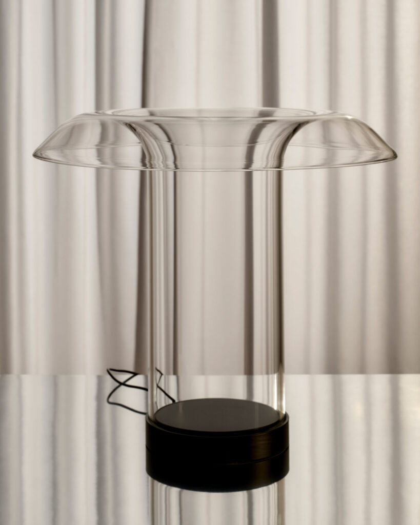 Minimal glass table lamp with a wide, прозрач diffuser and slim cylindrical body, resting on a black base with near-invisible light source
