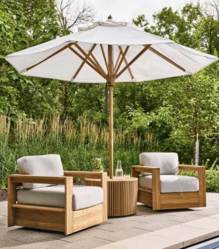 Outdoor lounge furniture with teak armchairs and a round side table sits beneath a wood-framed umbrella, creating a shaded, relaxed patio moment surrounded by soft planting.