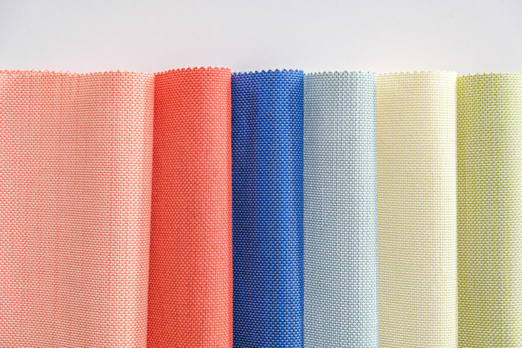 Sorbet-toned outdoor textiles in Batyline® Eden—rolled mesh fabric in peach, coral, denim blue, mint, and citrus hues showcasing woven texture and durability.
