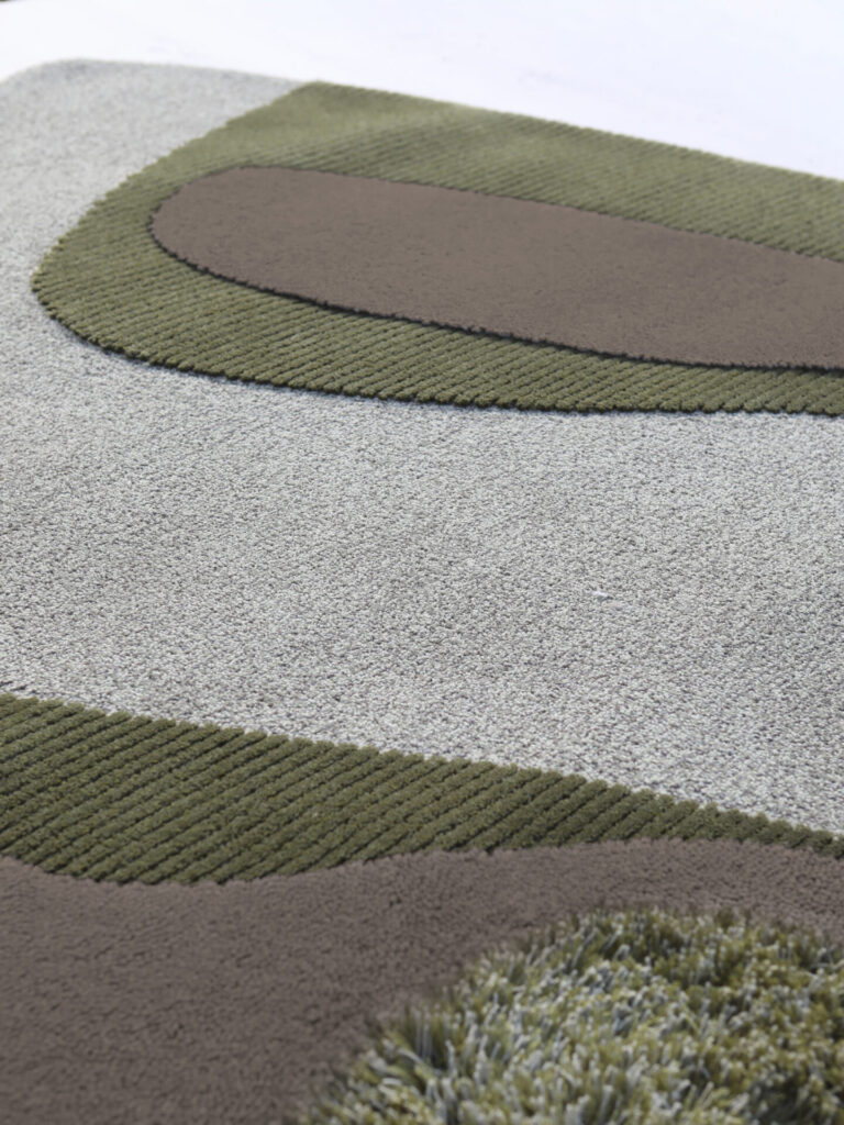 Close-up of organic area rugs showing textured pile variation in green and taupe, with sculpted edges and layered surfaces that evoke natural terrain.