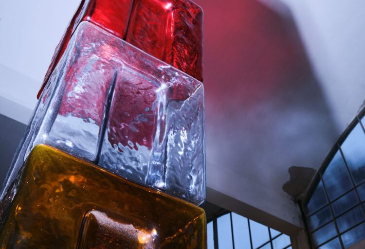 Towering stack of red, clear, and amber glass volumes forming a vertical glass lighting installation beneath curved industrial windows.