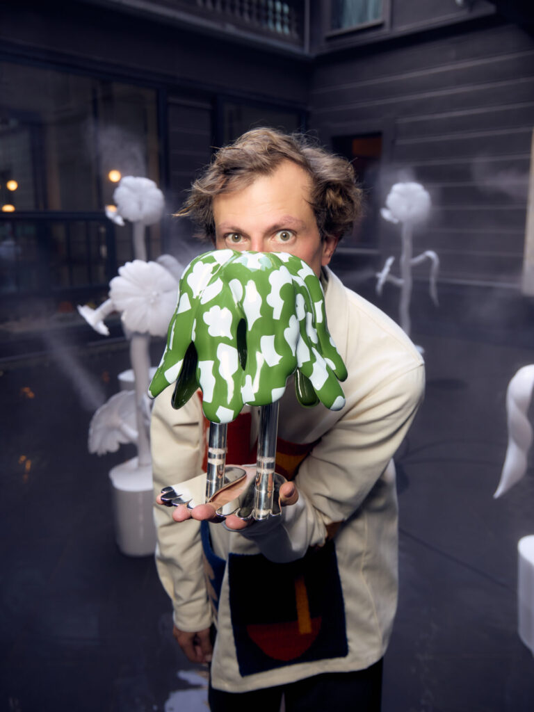 Matteo Cibic presents a small green and white glazed ceramic object with sculptural legs, set against the larger installation backdrop