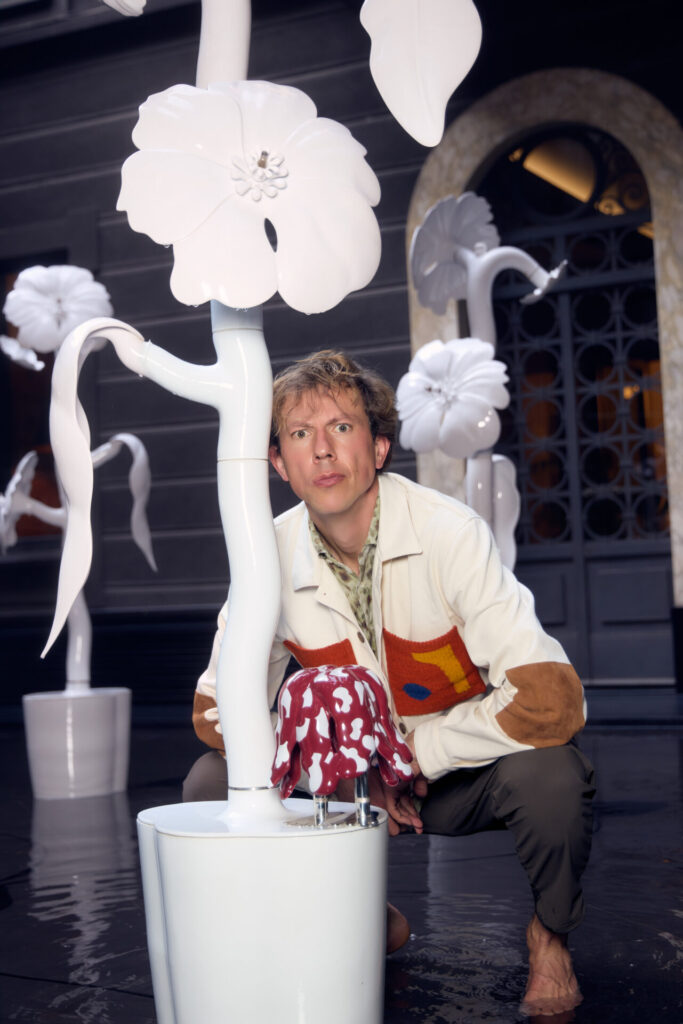 Designer Matteo Cibic crouches among large-scale ceramic flower sculptures, holding a playful glazed object within the installation