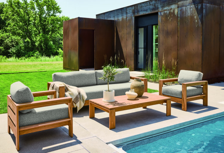 Outdoor lounge furniture in teak frames a poolside setting, with deep cushioned seating and a low table set against a weathered steel-clad structure and lush greenery.