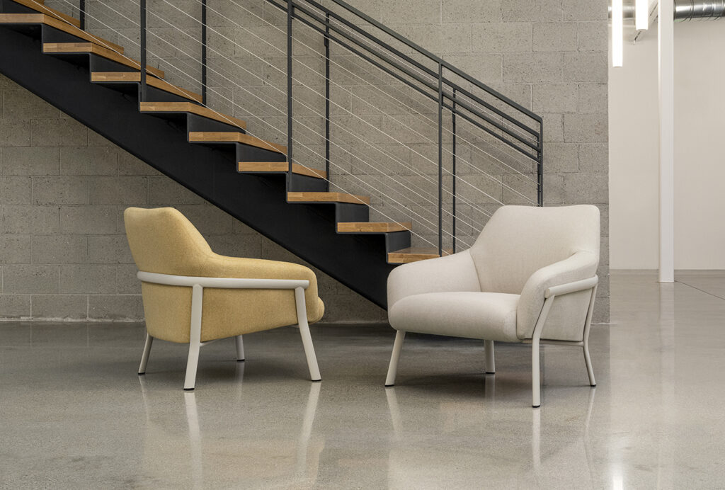 Pair of Talina lounge chairs in neutral and mustard tones beneath an industrial staircase, illustrating versatile contract lounge seating in open spaces.