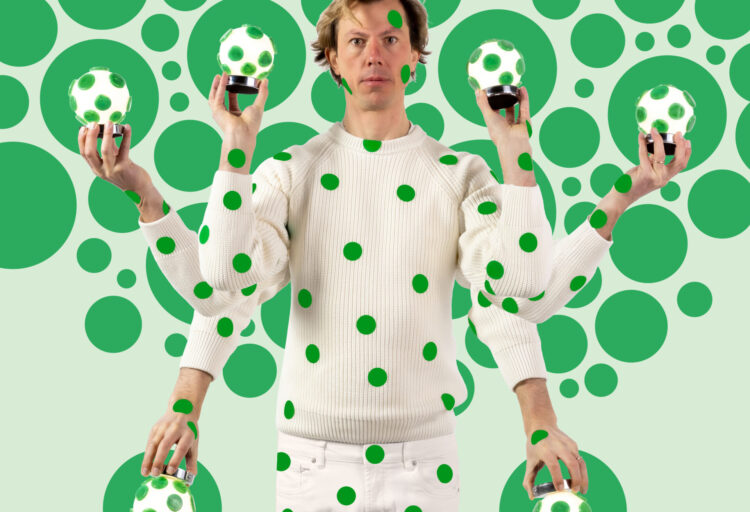 Designer Matteo Cibic poses against a field of oversized green polka dots while holding multiple glowing Tripop cordless table lamps in playful Murano glass with matching green spots.