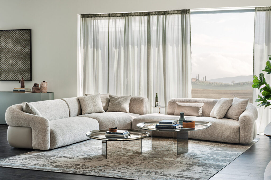 \Cattelan Italia’s Baldwin curved sofas in textured neutral upholstery create a modular conversation area with low metallic coffee tables and soft daylight filtering through linen curtains.