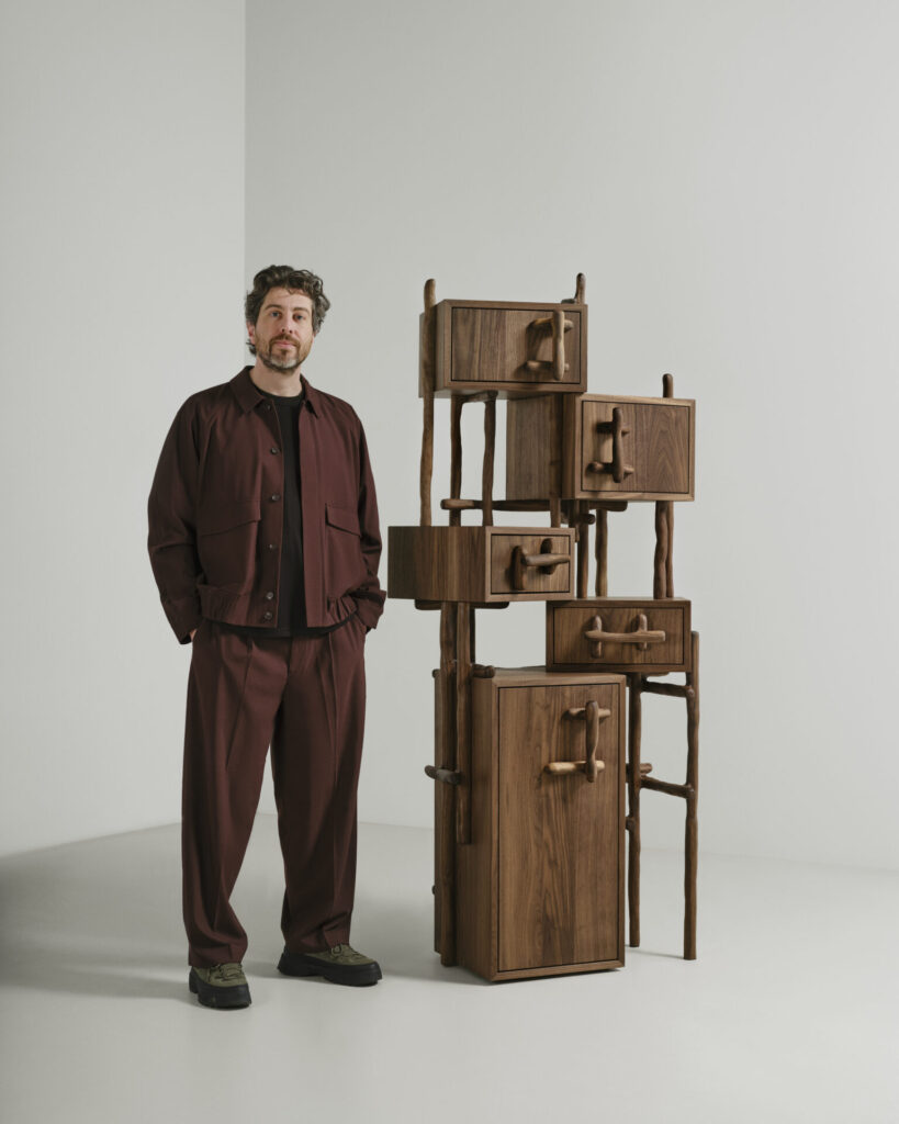 Designer Onno Adriaanse standing beside a sculptural walnut storage tower from the Hedera collection for Dilmos during Milan Design Week 2026.