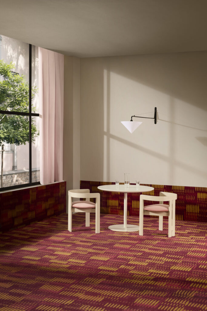 Burgundy and ochre patterned commercial carpet from Radici’s Pigments Collection adds painterly rhythm beneath pale pink curtains and café seating in a hospitality setting.