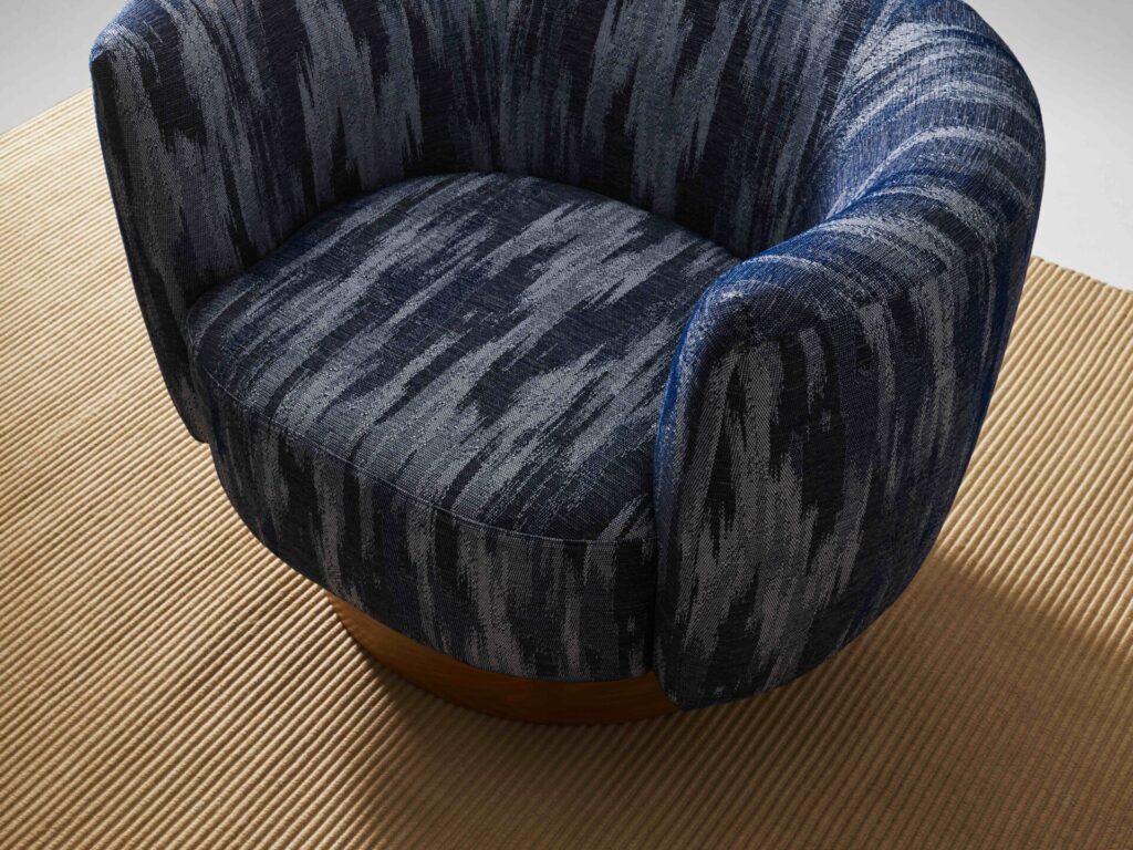 Hands hold an oversized olive-green cushion upholstered in Carnegie Xorel Luxe Lucia fabric, highlighting the soft woven texture and residential feel of the sustainable commercial upholstery.