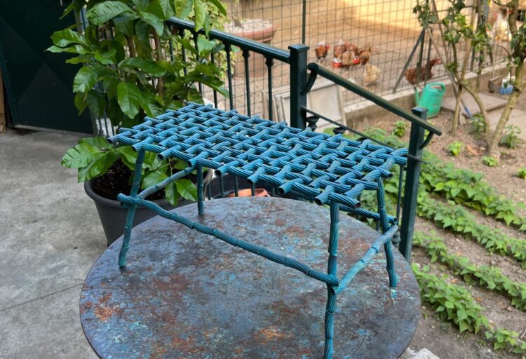 Cast bronze furniture stool by Chris Fusaro displayed outdoors, its woven tortiglioni structure casting a grid-like shadow across a weathered metal tabletop.