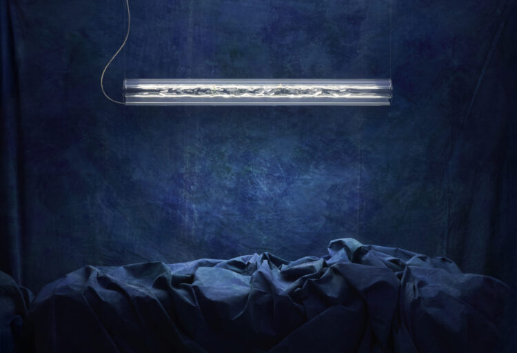 NALUM linear pendant lighting by Ingo Maurer hangs above draped blue fabric, its handcrafted rippled glass tube glowing like a frozen ocean wave.