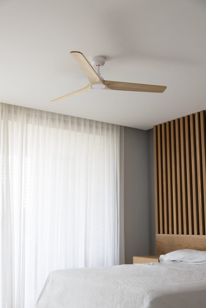 Faro Barcelona Klim architectural ceiling fan with light wood blades and white motor mounted above a minimalist bedroom with wood slat headboard and sheer curtains.