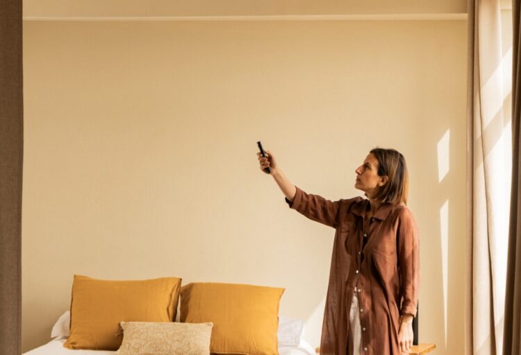 Faro Barcelona Klim architectural ceiling fan with light wood blades and white motor mounted above a minimalist bedroom with wood slat headboard and sheer curtains.