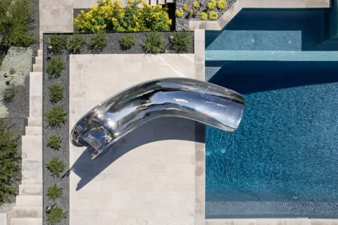 Aerial view of the Splinterworks Vertex stainless steel pool slide curving over a swimming pool.
