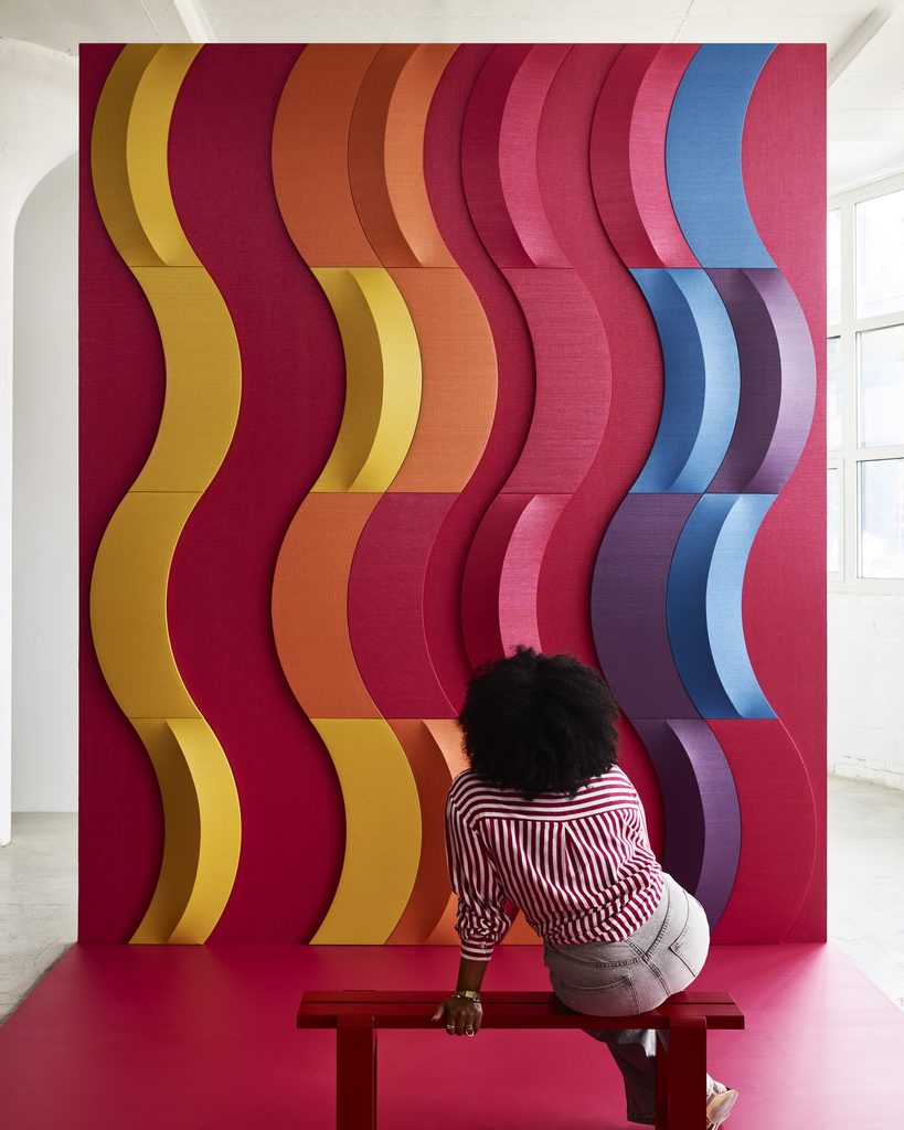 Multicolored Xorel Artform Bow acoustic wall tiles form vertical waves with raised crescent details behind a red bench.